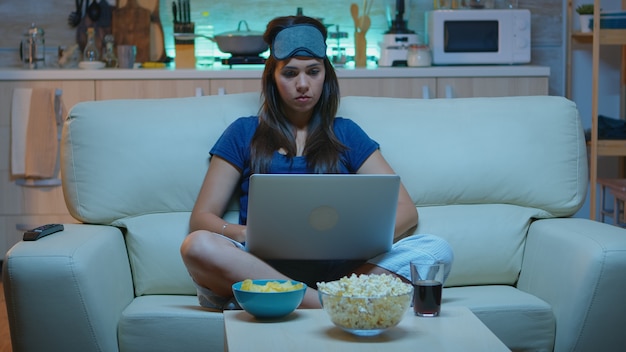 Señora en pijama y tapa de ojos en la frente escribiendo en la computadora portátil y viendo la televisión a altas horas de la noche. Freelancer trabajando sentado en el sofá leyendo escribiendo buscando navegar en un portátil usando tecnología de internet