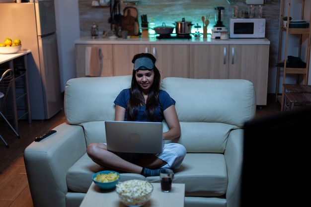 Señora en pijama y tapa de ojos en la frente escribiendo en la computadora portátil y viendo la televisión a altas horas de la noche. Freelancer trabajando sentado en el sofá leyendo escribiendo buscando navegar en un portátil usando tecnología de internet