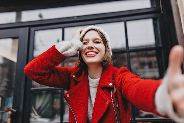 Señora positiva en abrigo rojo y gorro de punto se ríe con los ojos cerrados y hace selfie en la calle contra la ventana.