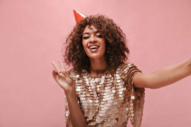 Señora positiva con cabello ondulado en gorra de vacaciones y vestido brillante que muestra el signo de la paz y toma una foto en la pared rosa aislada.