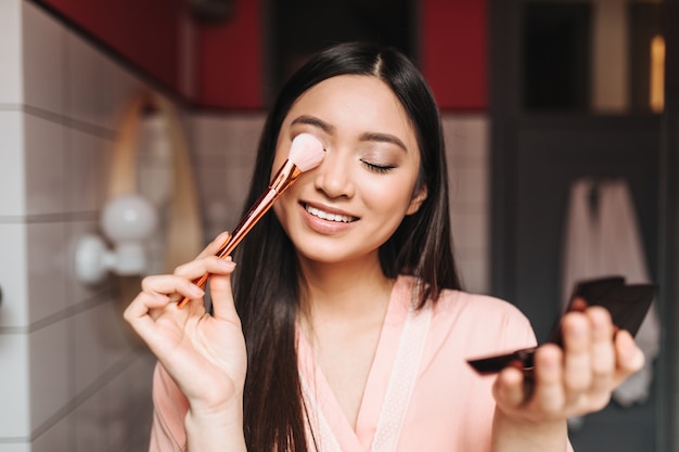 Señora positiva con cabello oscuro cubre sus ojos con pincel de maquillaje y posa en el baño.