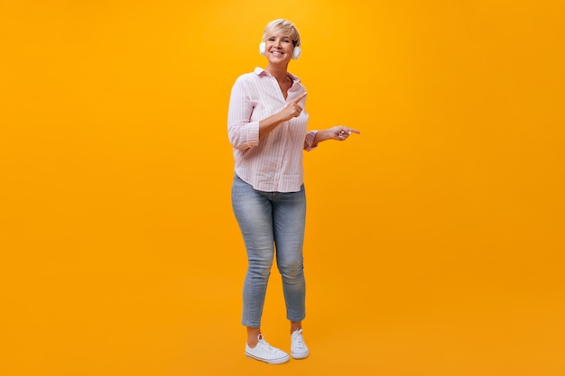 Señora positiva en jeans y camisa bailando y escuchando música en auriculares