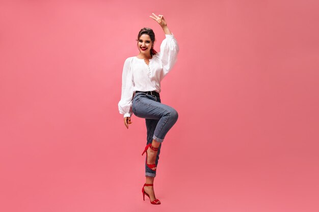 Señora positiva en traje de mezclilla muestra el signo de la paz sobre fondo rosa. Mujer joven feliz de buen humor en blusa blanca sonriendo.