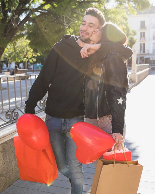 Señora que besa al individuo sonriente joven con los paquetes y el globo en la calle