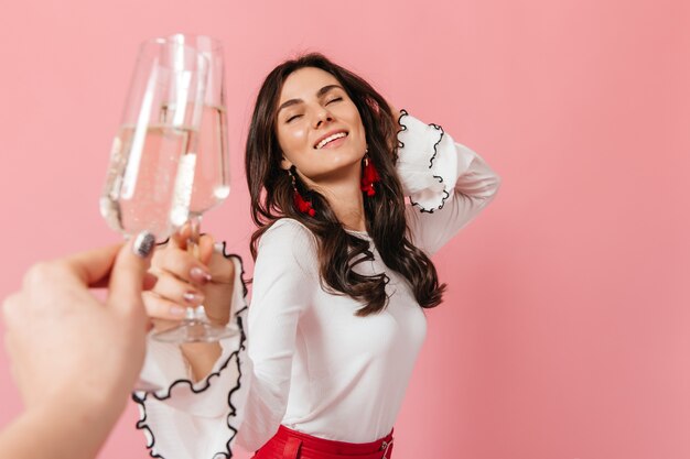 Señora rizada disfrutando de tintineo de copas de vino. Retrato de niña sonriendo con los ojos cerrados sobre fondo rosa.