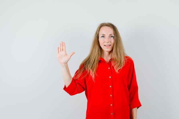 Señora rubia agitando la mano para decir adiós en camisa roja y mirando confiada.