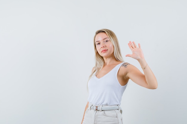 Señora rubia agitando la mano para decir adiós en camiseta, pantalones y luciendo confiada