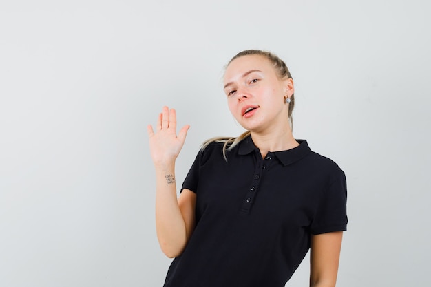 Señora rubia agitando la mano para saludar en camiseta negra y mirando confiada, vista frontal.