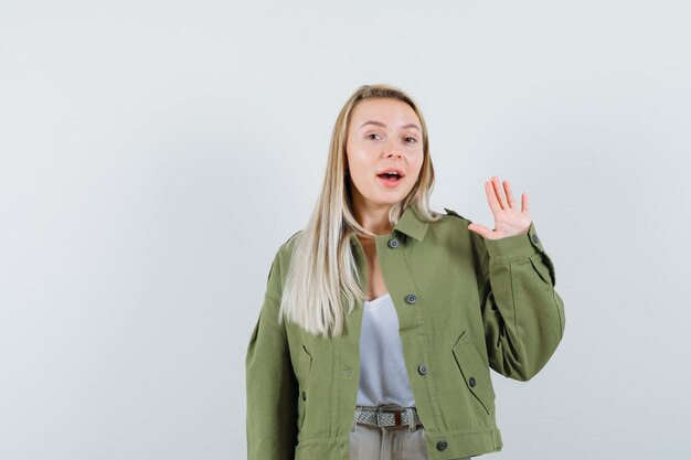 Señora rubia agitando la mano para saludar en chaqueta, pantalón y mirando alegre, vista frontal.