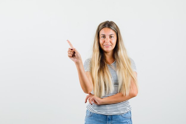 Señora rubia apuntando hacia afuera en camiseta y mirando seguro, vista frontal.