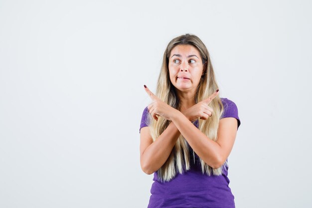 Señora rubia apuntando hacia afuera con camiseta violeta y mirando confundida, vista frontal.