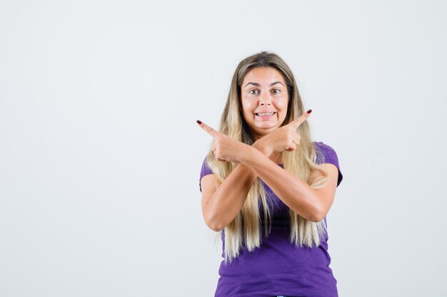 Señora rubia apuntando hacia afuera en camiseta violeta y mirando emocionada, vista frontal.