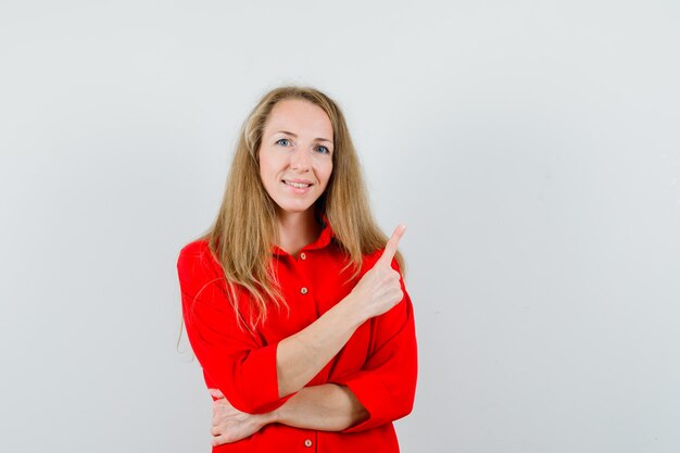 Señora rubia apuntando hacia arriba con camisa roja y mirando feliz.