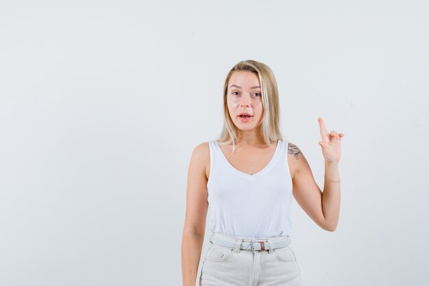 Señora rubia apuntando hacia arriba en camiseta, pantalones y mirando confiada