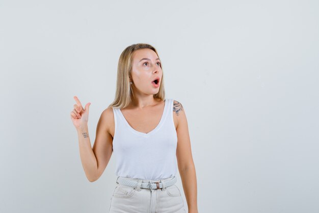 Señora rubia apuntando hacia arriba en camiseta, pantalones y mirando sorprendido, vista frontal.