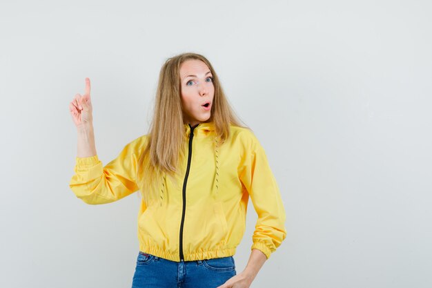 Señora rubia apuntando hacia arriba en chaqueta, jeans y mirando confiada.