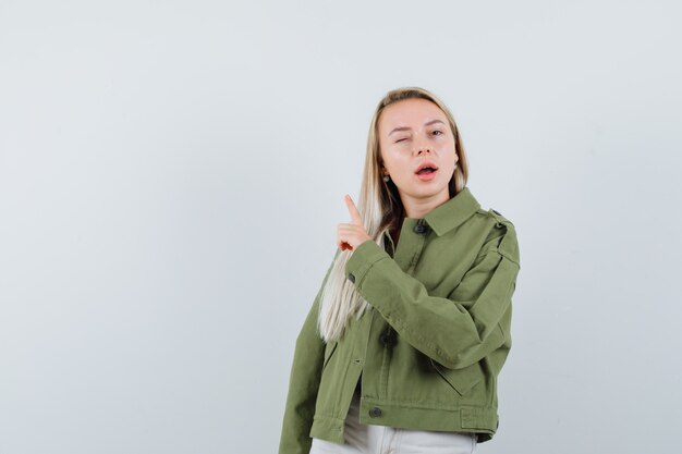 Señora rubia apuntando hacia arriba en chaqueta, pantalones y mirando confiada, vista frontal.