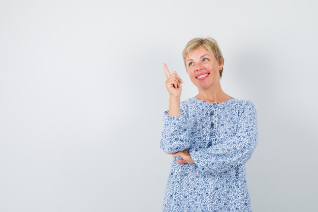 Señora rubia apuntando hacia arriba mientras sonríe con blusa estampada y luce alegre, vista frontal. espacio para texto