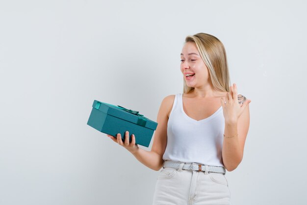 Señora rubia apuntando hacia arriba, sosteniendo la caja actual en camiseta, pantalones y mirando feliz, vista frontal.