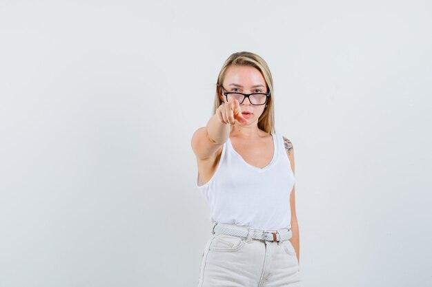 Señora rubia apuntando a la cámara en camiseta, pantalones y mirando confiada. vista frontal.