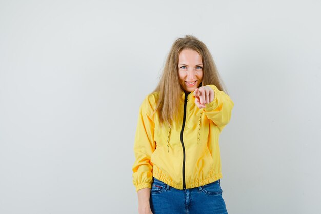 Señora rubia apuntando a la cámara en chaqueta, jeans y mirando alegre,