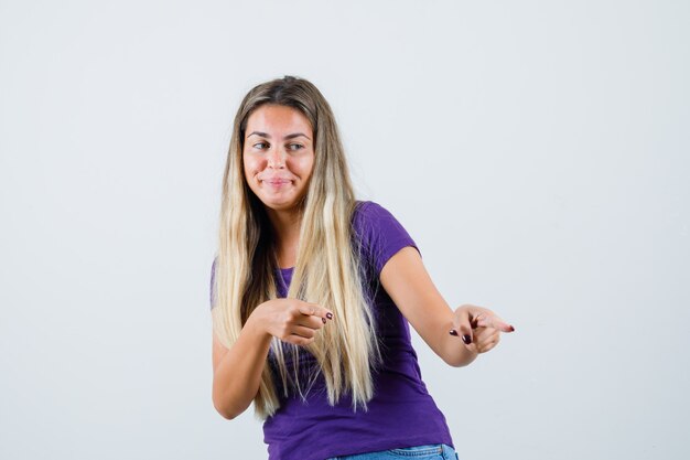 Señora rubia apuntando con camiseta violeta, jeans y mirando alegre, vista frontal.