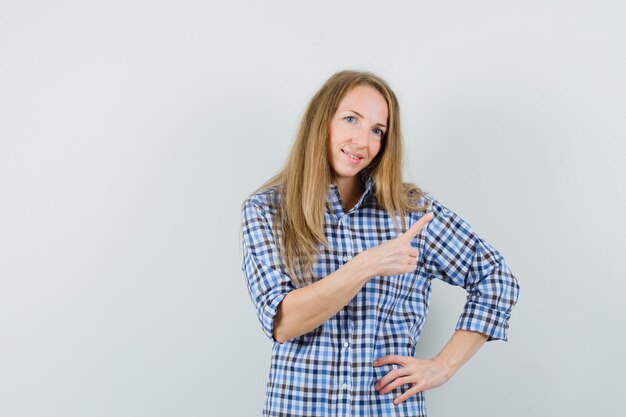 Señora rubia apuntando a la esquina superior derecha en camisa y mirando alegre.