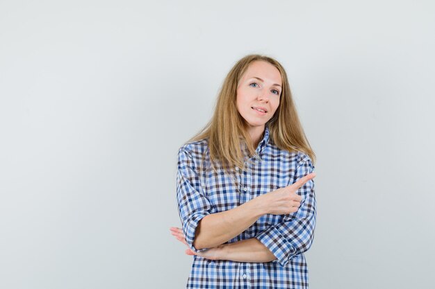 Señora rubia apuntando a la esquina superior derecha en camisa y mirando confiada,