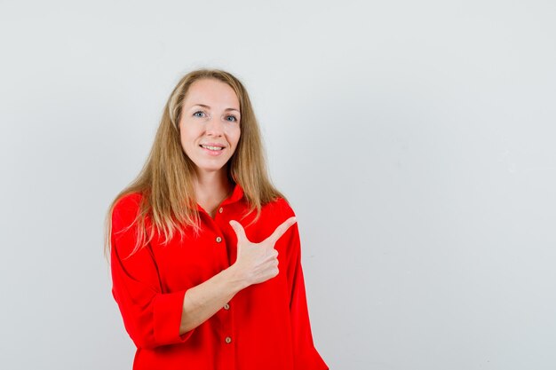 Señora rubia apuntando a la esquina superior derecha con camisa roja y mirando alegre.