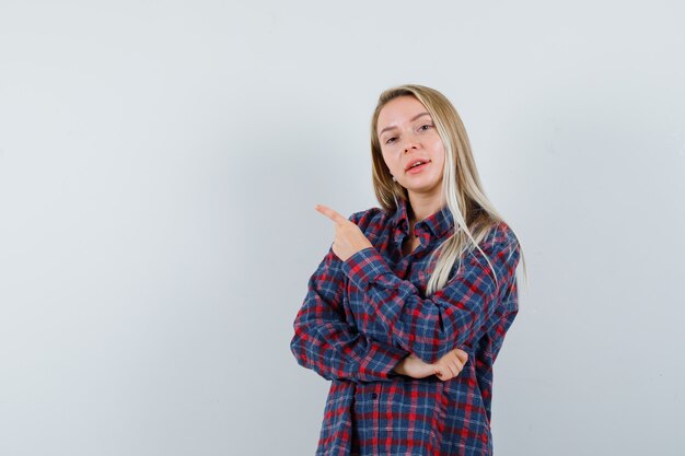 Señora rubia apuntando a la esquina superior izquierda en camisa casual y mirando confiada, vista frontal.