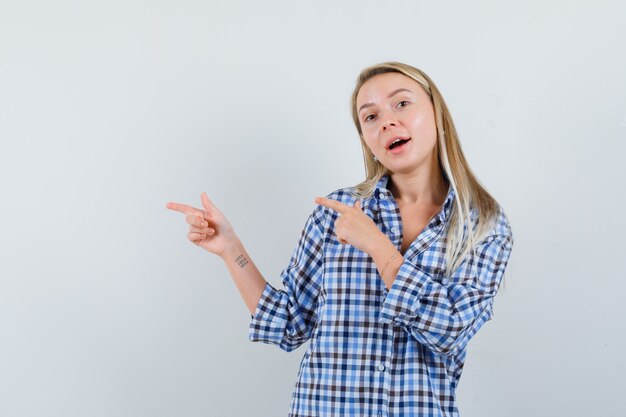 Señora rubia apuntando a un lado en camisa casual y mirando alegre