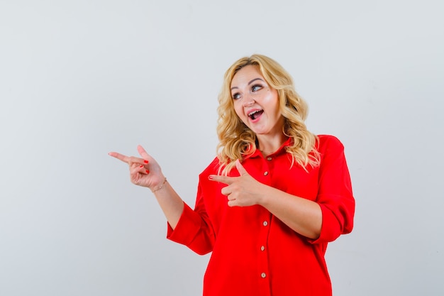 Señora rubia apuntando a un lado en camisa roja y mirando alegre.