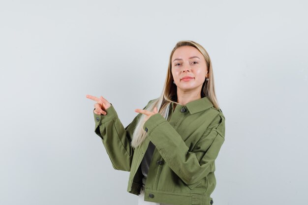 Señora rubia apuntando a un lado en chaqueta, pantalones y mirando confiada, vista frontal.