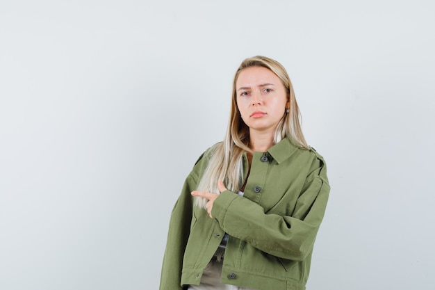 Señora rubia apuntando a un lado en chaqueta, pantalones y mirando insatisfecho, vista frontal.