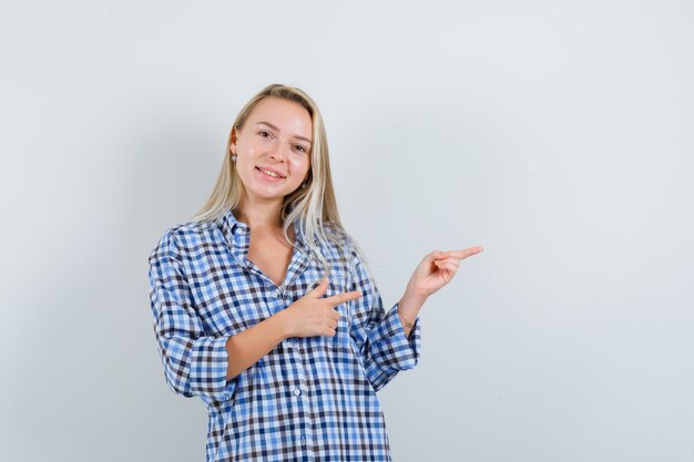 Señora rubia apuntando hacia el lado derecho con camisa a cuadros y mirando alegre