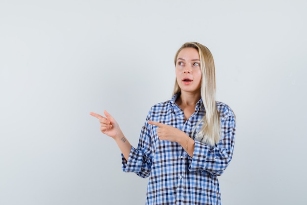 Señora rubia apuntando hacia el lado izquierdo con camisa a cuadros y mirando asombrado