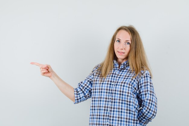 Señora rubia apuntando hacia el lado izquierdo en camisa y mirando confiada,