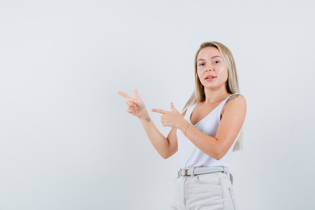 Señora rubia apuntando hacia el lado izquierdo en camiseta, pantalones y luciendo confiada