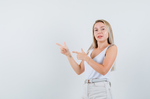 Señora rubia apuntando hacia el lado izquierdo en camiseta, pantalones y luciendo confiada