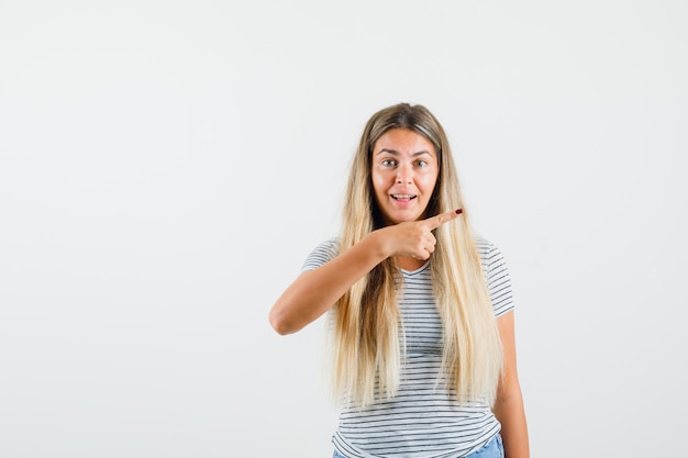 Señora rubia apuntando hacia otro lado en camiseta y mirando divertido. vista frontal.