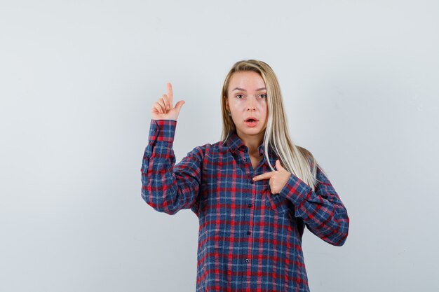 Señora rubia apuntando a sí misma y con camisa casual y mirando maravillada, vista frontal.