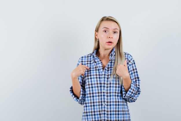 Señora rubia apuntando a sí misma en camisa a cuadros y mirando confundida