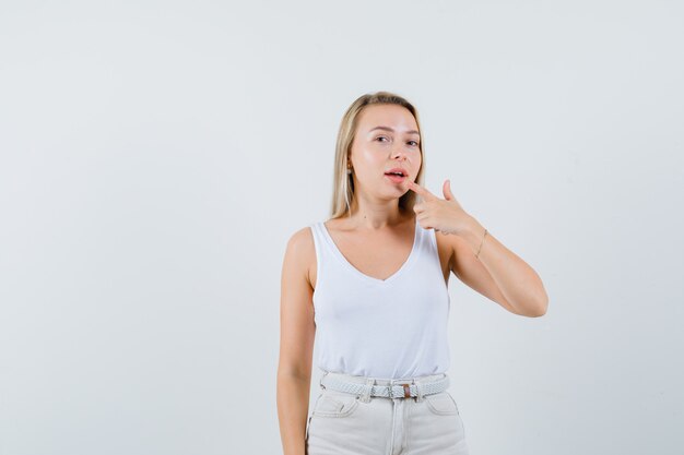 Señora rubia apuntando a su boca en camiseta, pantalones y luciendo linda