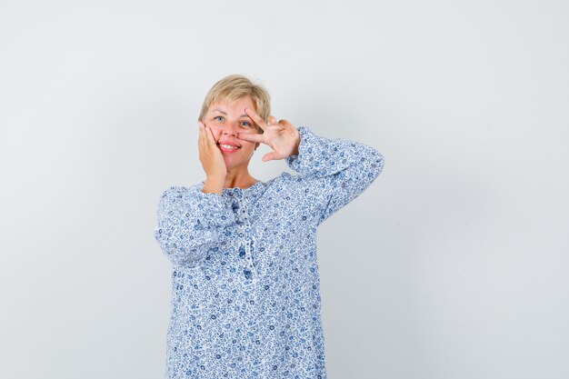 Señora rubia en blusa estampada que muestra el signo v sobre su ojo y luce alegre, vista frontal.