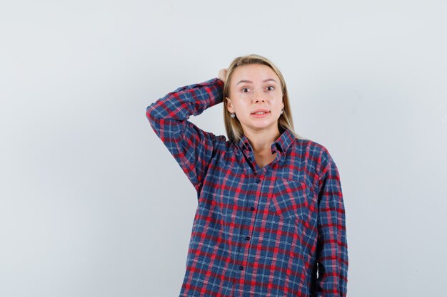 Señora rubia en camisa casual manteniendo la mano en la cabeza y mirando olvidadizo, vista frontal.