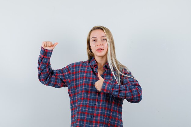 Señora rubia en camisa casual mostrando el pulgar hacia arriba, apuntando hacia otro lado y mirando confiado, vista frontal.