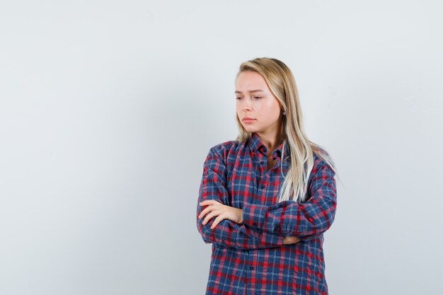 Señora rubia en camisa casual de pie con los brazos cruzados y mirando triste, vista frontal.