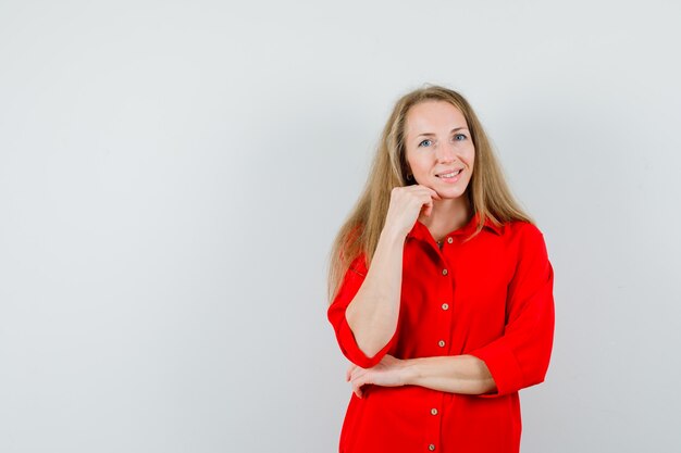 Señora rubia en camisa roja de pie en pose de pensamiento y mirando alegre,