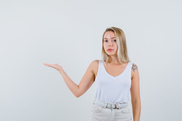 Señora rubia en camiseta, pantalones dando la bienvenida o mostrando algo y luciendo confiada