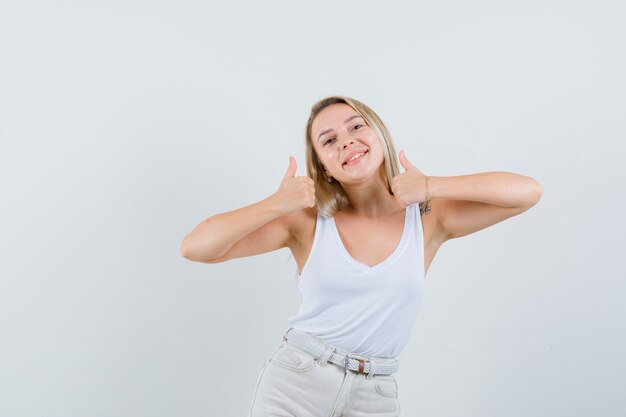 Señora rubia en camiseta, pantalones mostrando el pulgar doble hacia arriba y mirando alegre, vista frontal.